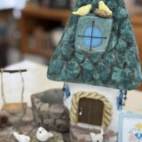 青緑色のとんがり屋根が特徴の小さな家をモルタルで作った作品。家の前には白いフェンスやブランコ、石の上の小鳥たち、玄関横には小さな猫の姿もあり、細部まで丁寧に作り込まれている。童話の世界のような温かく可愛らしい雰囲気が漂う。