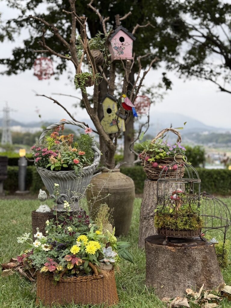 庭の芝生の上に、花や多肉植物を植えたバスケットや鉢が並ぶナチュラルなガーデンディスプレイ。切り株や古いカゴ、鳥かごを使った寄せ植えが配置され、背景の木には色とりどりの鳥の巣箱や小物が飾られています。自然の中に手作りの温かみが感じられる、可愛らしい庭の一角です。