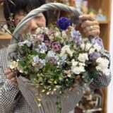 女性が両手で大きな花かごを持ち、笑顔で顔をのぞかせている。かごには紫や白、淡いピンクの花々がたっぷりと生けられ、葉の緑と調和して華やか。背景には木製の棚があり、温かみのある室内の雰囲気が感じられる。