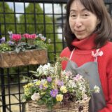 赤いセーターにグレーのエプロンを着た女性が、花かごを抱えて微笑んでいる。かごには黄色、白、紫などの小花が咲き、明るく華やかな寄せ植え。背景には黒いフェンスと緑の植木があり、屋外の爽やかな雰囲気が感じられる。