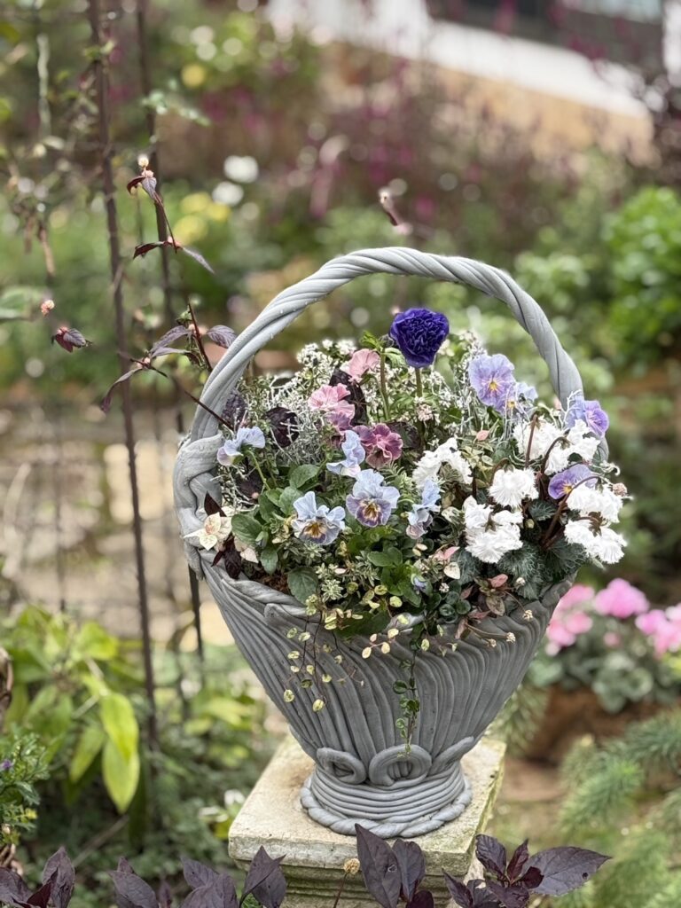 庭の一角に置かれたグレーの花かごに、紫や白、淡いピンクの花々が美しく咲いている。細かな葉やつるが垂れ下がり、ナチュラルで上品な寄せ植え。背景には緑の植物や花がぼんやりと広がり、穏やかな庭の雰囲気が漂っている。