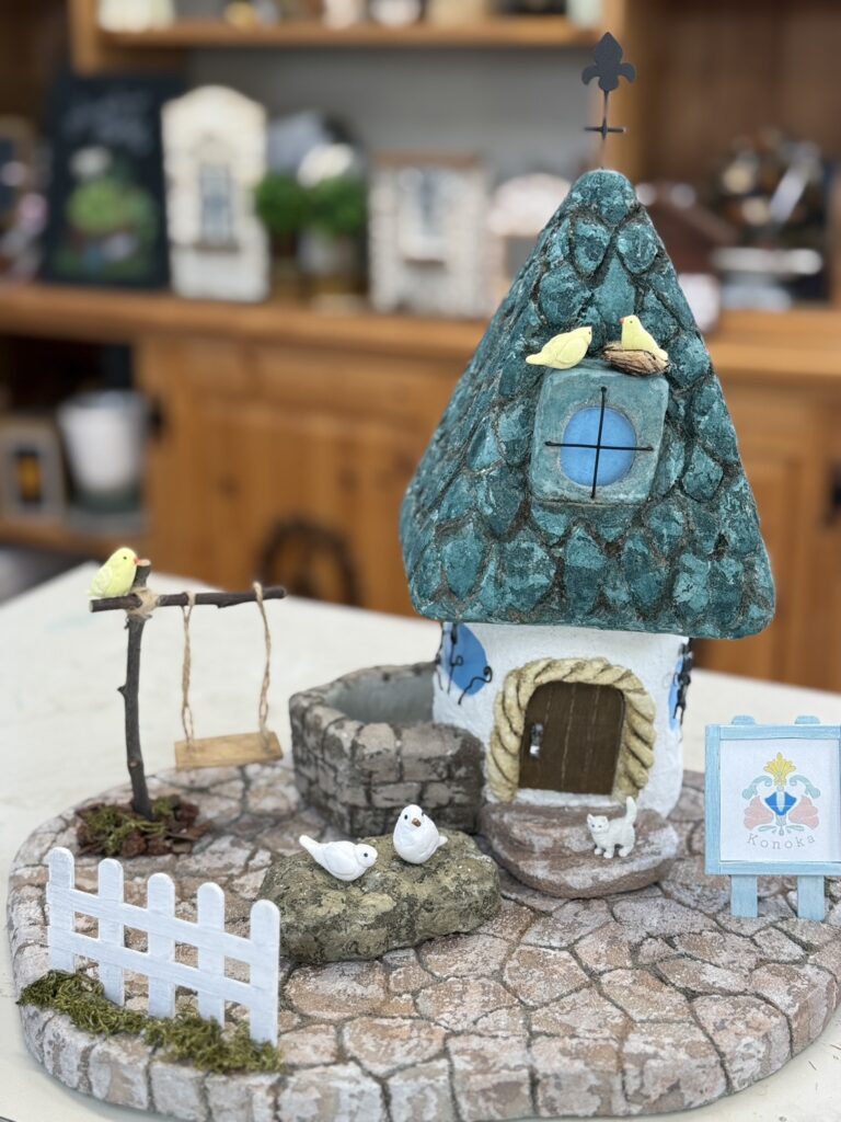 青緑色のとんがり屋根が特徴の小さな家をモルタルで作った作品。家の前には白いフェンスやブランコ、石の上の小鳥たち、玄関横には小さな猫の姿もあり、細部まで丁寧に作り込まれている。童話の世界のような温かく可愛らしい雰囲気が漂う。