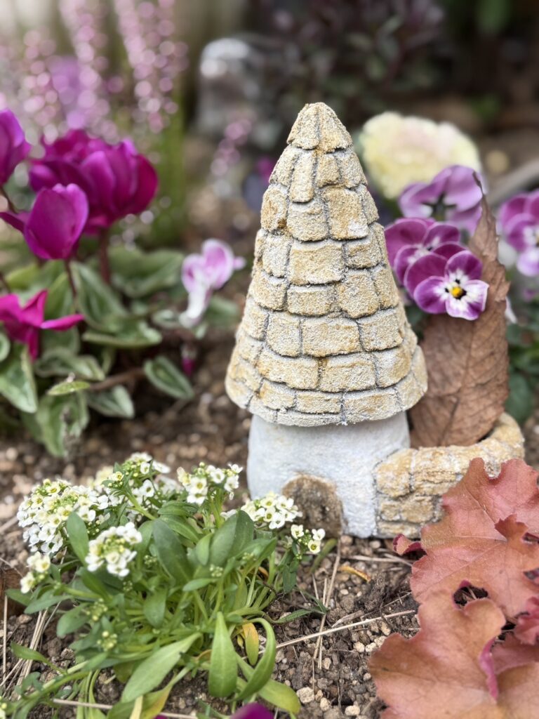 花壇の土の上に置かれた、モルタルで作られた小さなコビトハウスのアート作品。石を積み重ねたような質感の円すい形の屋根と、白っぽい壁に小さな入口がある。周囲には紫や白の花、茶色く色づいた葉が植えられており、物語の世界のような静かであたたかい雰囲気が感じられる。