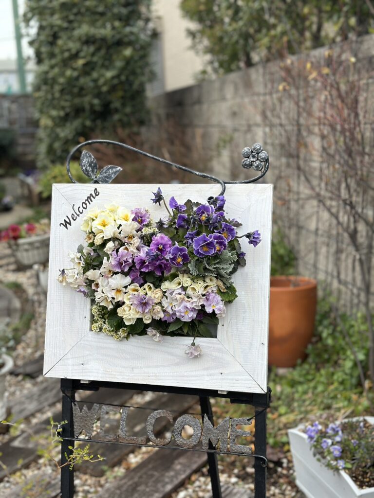 白いフレーム型のウェルカムボードに、紫とクリーム色のビオラを立体的に寄せ植えしたアレンジメント。上部には黒いアイアンの葉付き装飾、下のスタンドには「WELCOME」の文字。背景は庭の緑とレンガ塀で、屋外に設置された華やかな作品が映えている。
