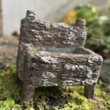 古びた木箱のような質感を持つ小さな容器が、苔の生えた地面の上に置かれている様子。表面はざらつき、白い風化跡がまだらに残っていて、脚がついた四角い形。背景にはぼんやりと緑の植物が見え、全体的に自然の中に溶け込んだような雰囲気。