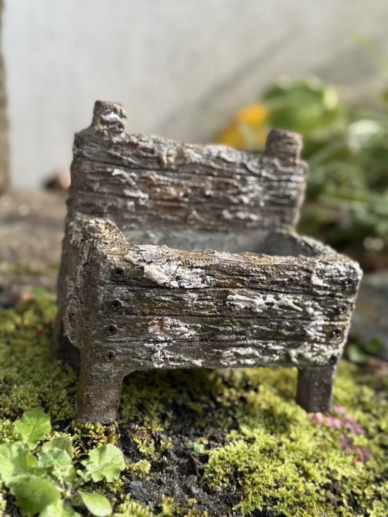 古びた木箱のような質感を持つ小さな容器が、苔の生えた地面の上に置かれている様子。表面はざらつき、白い風化跡がまだらに残っていて、脚がついた四角い形。背景にはぼんやりと緑の植物が見え、全体的に自然の中に溶け込んだような雰囲気。