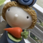 白い柵にもたれかかるように置かれた、うさぎと子どもを組み合わせたようなミニチュア人形。青い服を着た丸い顔の人形が、オレンジ色のにんじんを両手で抱えている。頭の上には青いうさぎの顔と長い耳があり、全体に素朴でやさしい表情。室内に飾られた、かわいらしい雰囲気の置物。