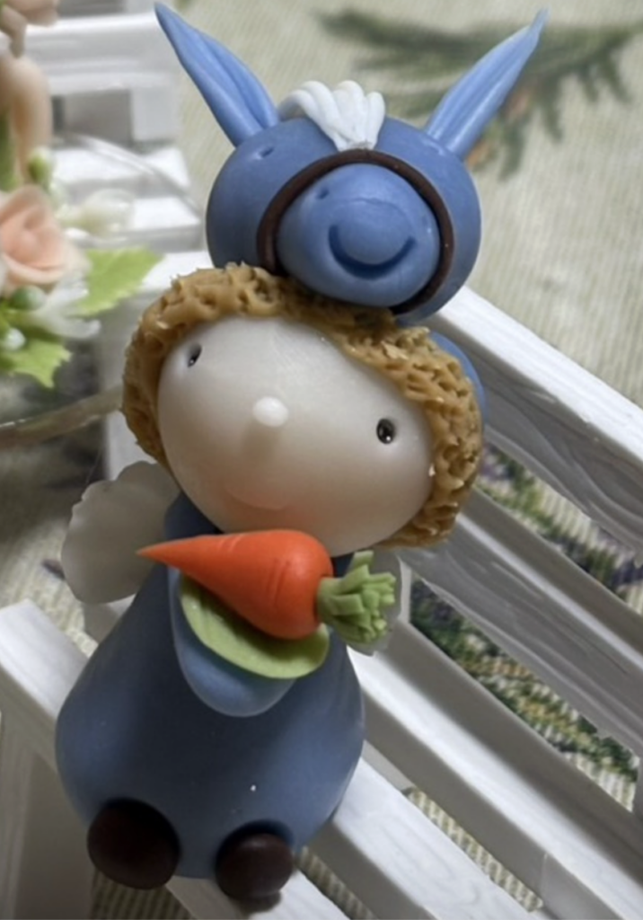 白い柵にもたれかかるように置かれた、うさぎと子どもを組み合わせたようなミニチュア人形。青い服を着た丸い顔の人形が、オレンジ色のにんじんを両手で抱えている。頭の上には青いうさぎの顔と長い耳があり、全体に素朴でやさしい表情。室内に飾られた、かわいらしい雰囲気の置物。