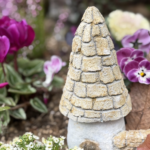 花壇の土の上に置かれた、モルタルで作られた小さなコビトハウスのアート作品。石を積み重ねたような質感の円すい形の屋根と、白っぽい壁に小さな入口がある。周囲には紫や白の花、茶色く色づいた葉が植えられており、物語の世界のような静かであたたかい雰囲気が感じられる。