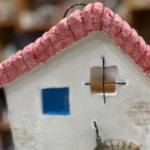 白いモルタルで作られた小さな家のアート作品。赤い瓦屋根に、青い四角い窓と十字格子の小窓、緑がかった扉がある。家の前には小さな自転車と三羽の白いアヒルの置物が並び、素朴で物語性のある雰囲気。背景は室内の棚で、作品にピントが合っている。