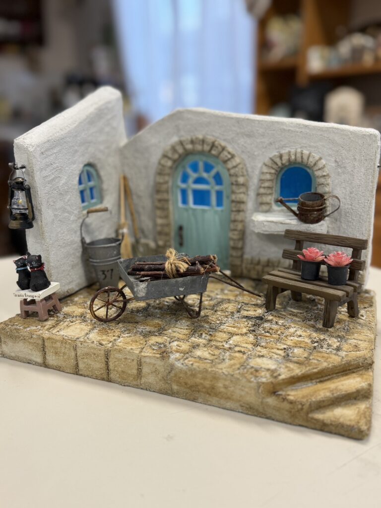 白い家の形をしたモルタルアート作品の置物。赤みのある瓦屋根と、ざらりとした白い壁が手作りらしい質感で表現されている。正面には青い小窓と、アーチ状の枠に囲まれたくすんだ緑色のドアがあり、上には小さなランプが付いている。足元には番号札の付いたミニチュアの三輪自転車と、並んで歩く白いアヒルが3羽置かれ、物語の一場面のような温かく可愛らしい雰囲気が伝わる。