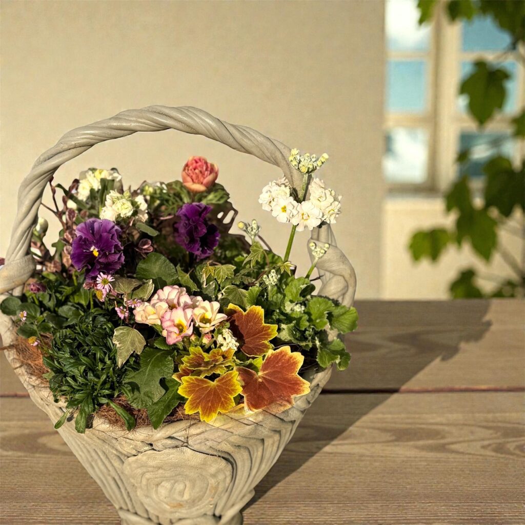 日差しの差し込む屋外の木製テーブルの上に置かれた、淡いグレーの取っ手付きバスケットの寄せ植え。紫やピンク、白の花と、緑や茶色がかった葉がバランスよく植えられ、春のやさしい雰囲気を感じさせる。