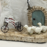 白い壁の小さな家をかたどったモルタルアート作品の一部を写したクローズアップ。アーチ状の石枠に囲まれた青緑色のドアの前に、番号「58」の札が付いたミニチュアの三輪自転車が置かれている。その横には、くちばしを少し開けた白いアヒルの置物が3羽、列になって立っている。床は石畳風で、全体に素朴で温かみのある、物語を感じさせる雰囲気が伝わる。