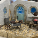 白い家の形をしたモルタルアート作品の置物。赤みのある瓦屋根と、ざらりとした白い壁が手作りらしい質感で表現されている。正面には青い小窓と、アーチ状の枠に囲まれたくすんだ緑色のドアがあり、上には小さなランプが付いている。足元には番号札の付いたミニチュアの三輪自転車と、並んで歩く白いアヒルが3羽置かれ、物語の一場面のような温かく可愛らしい雰囲気が伝わる。