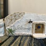 白い漆喰風のミニチュアハウスと猫の置物。「Welcome」の文字が入ったナチュラルなガーデン雑貨で、エス・オフィスガーデンの世界観を表現している。