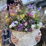 ピンクや紫、黄色の花々をバランスよく寄せ植えしたナチュラルなバスケット型鉢を両手で持つ女性。屋外で撮影された春の寄せ植え作品。