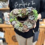 グリーンやピンク系の多肉植物をぎっしりと植え込んだハート型の多肉リースを両手で持つ女性。教室内で撮影された完成作品の様子。