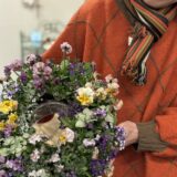 春色のビオラや小花で作られたリースを手に持つ女性参加者。レッスン会場で完成作品を披露している様子。