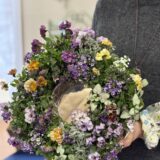 紫や淡い黄色のビオラ、小花やシルバーリーフをバランスよくあしらった春のフラワーリースを両手で持つ女性。教室で撮影された完成作品。