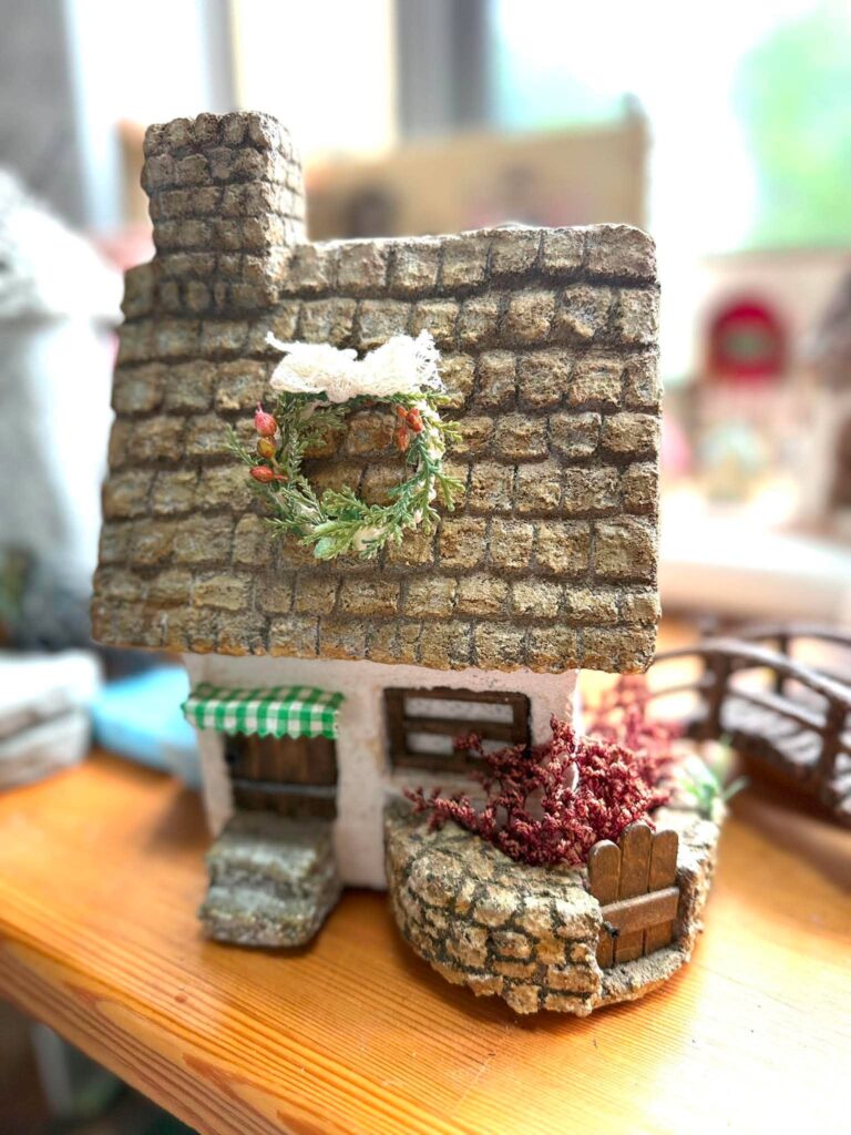 石積み風の屋根に緑のリースが飾られた、庭付きの小さなミニチュアハウス